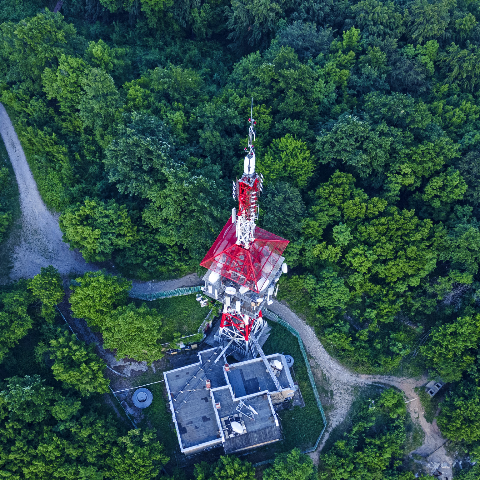 From above, a red telecommunications tower can be seen amidst a lush green forest.