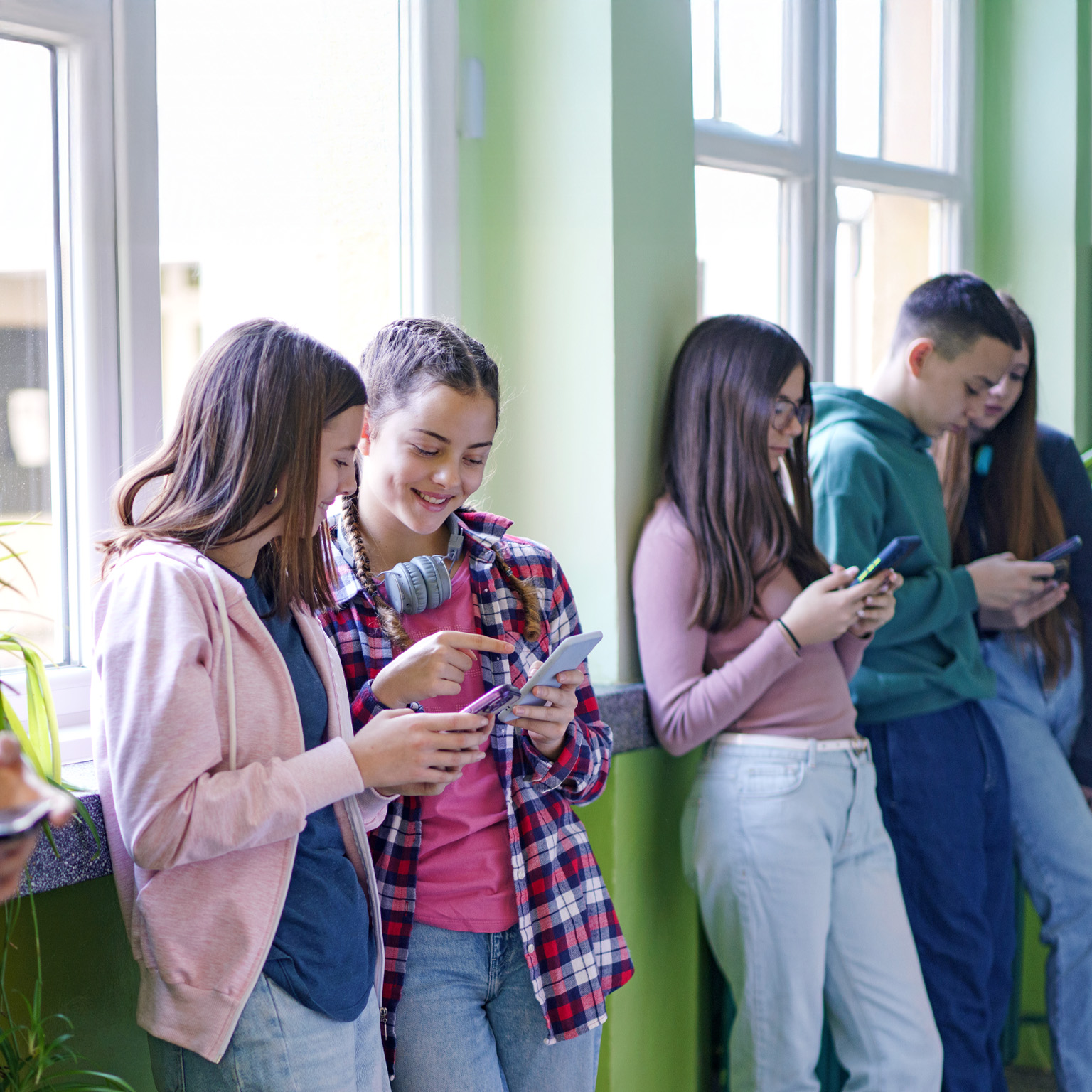 A group of teenagers are standing against a wall, looking at their smartphones. One of the girls in the middle is pointing at her phone, sharing something with another girl beside her.
