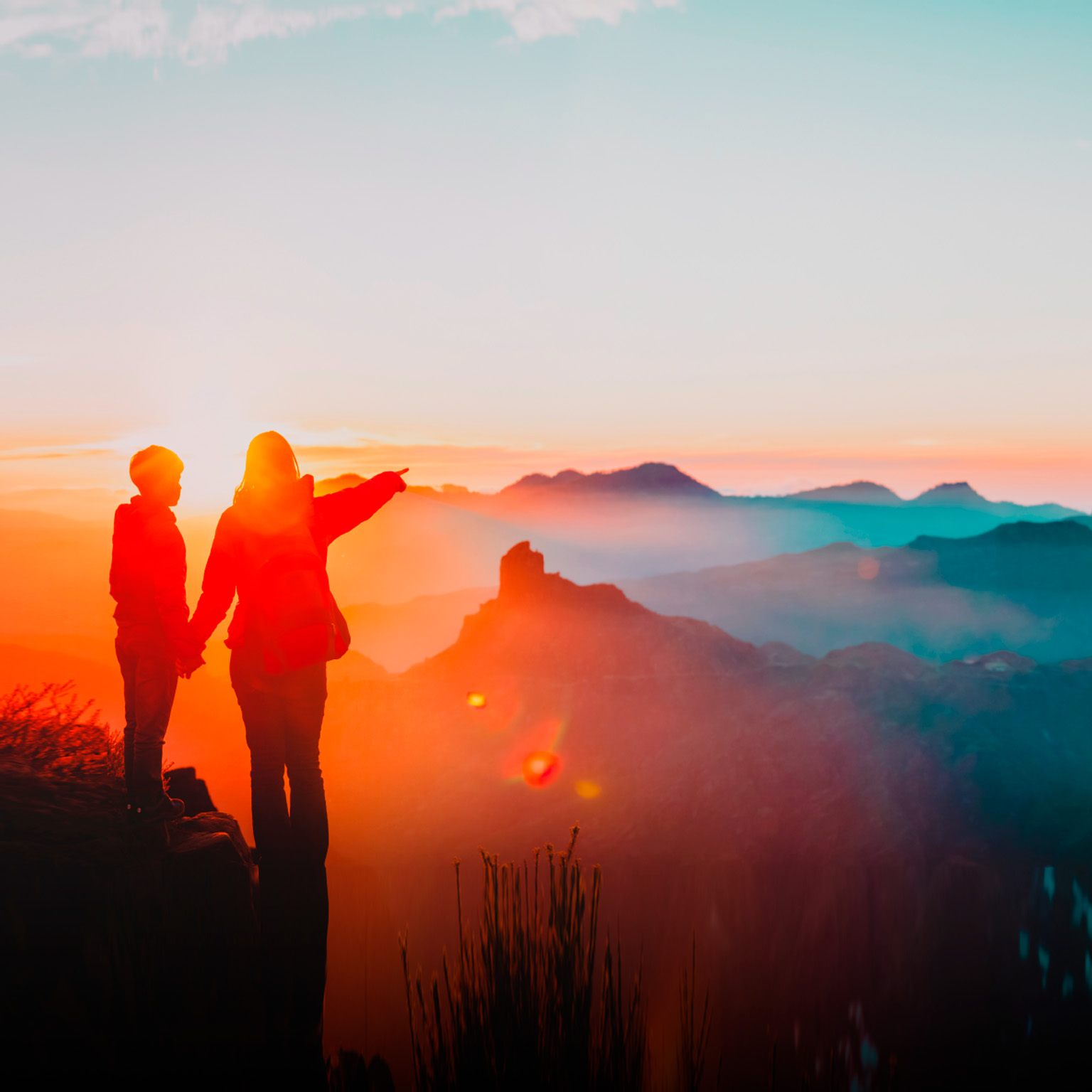 mother holding hand of son while pointing over canyon rim, sun setting in background - photo