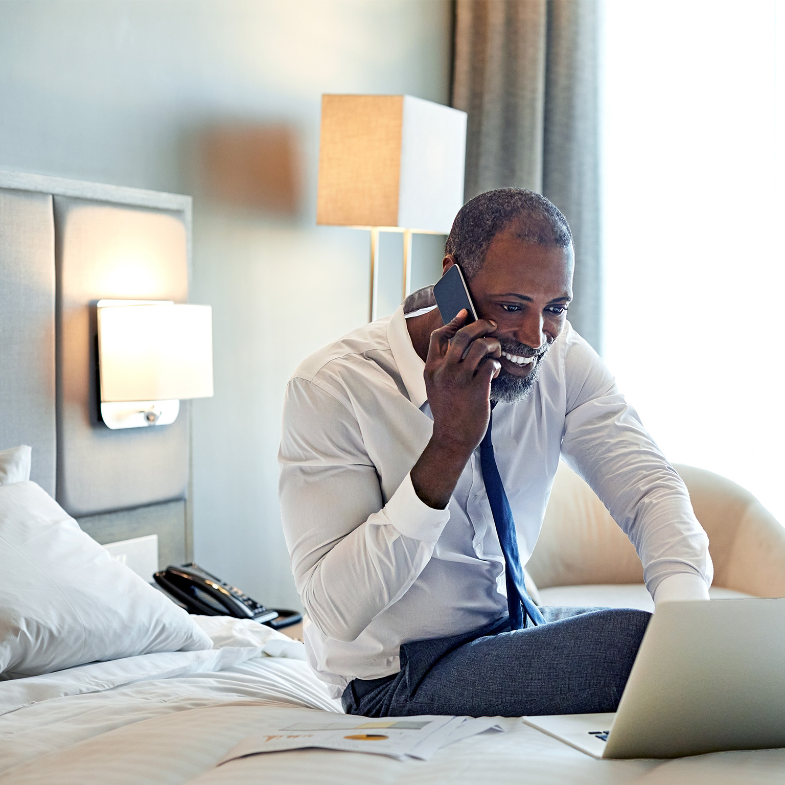 Male entrepreneur sitting on bed in a hotel room working on this laptop while on his cellphone