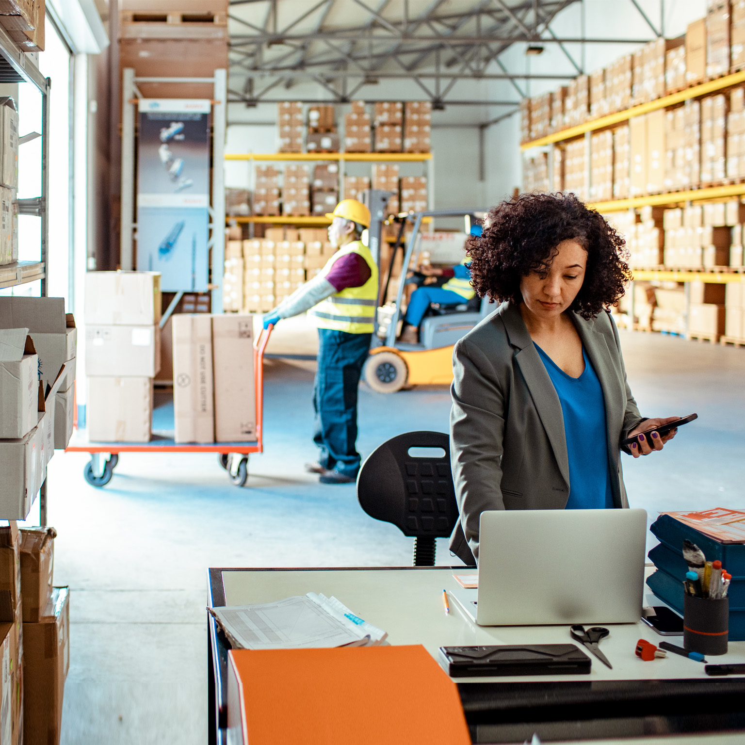 photo warehouse with woman looking at laptop while men move boxes in background