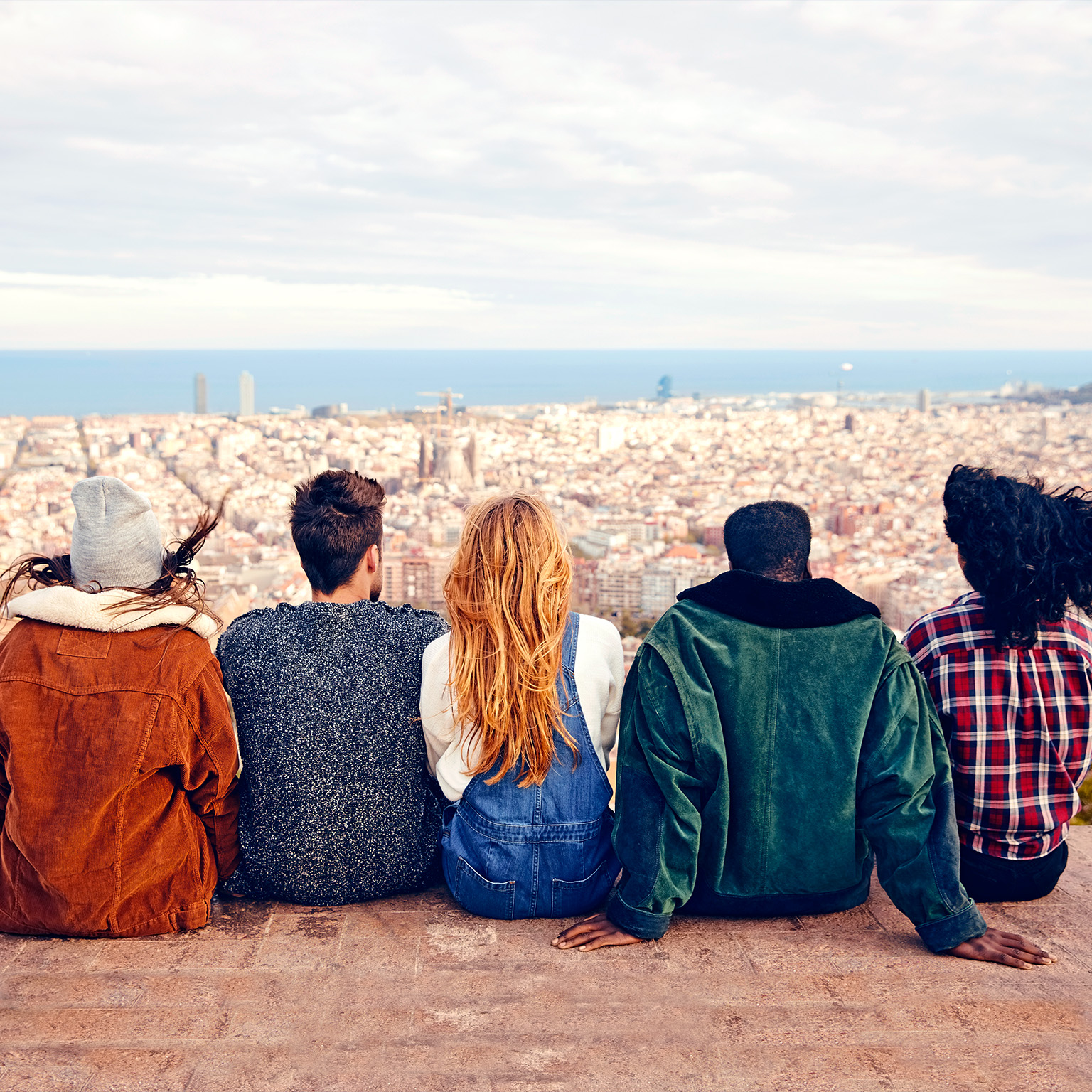 Image from behind of a group of friends sitting on a terrace looking out over a city with the ocean in the far distance.