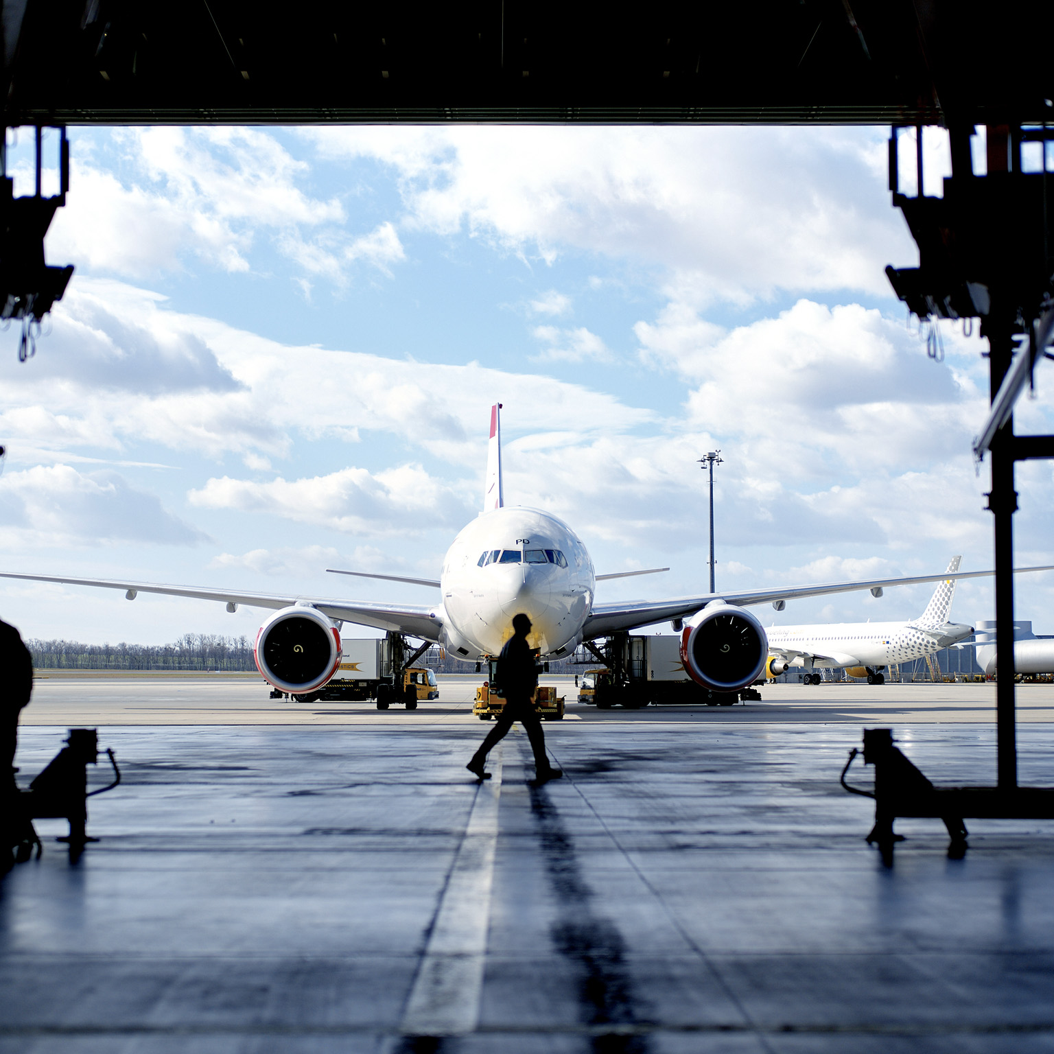 A large commercial airplane parked on the tarmac, viewed from inside a hangar with silhouetted workers and equipment framing the scene. A person is walking through the foreground while the bright sky and clouds create a striking contrast behind the aircraft.