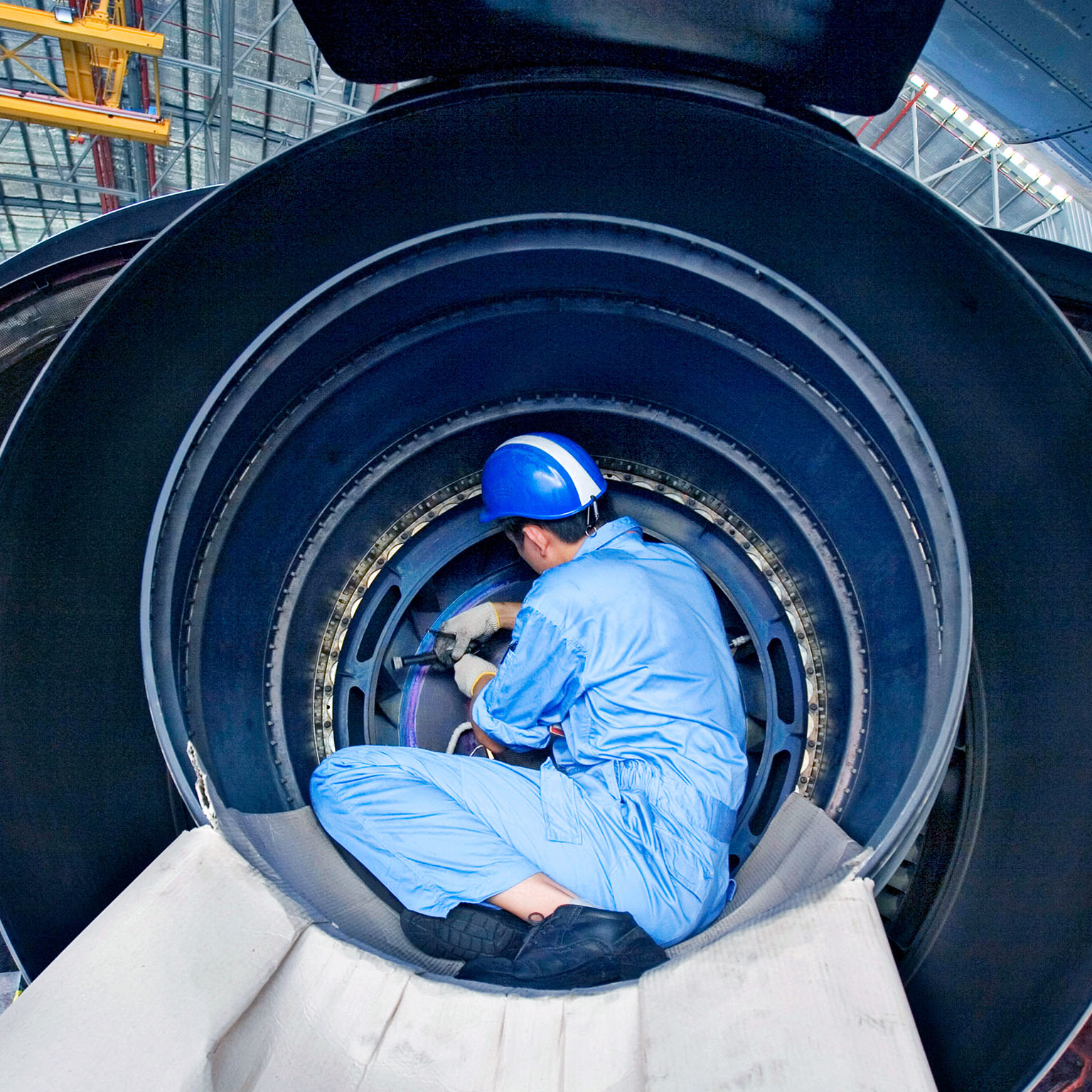 A maintenance technician in protective gear works inside a large jet engine in an aircraft hangar.