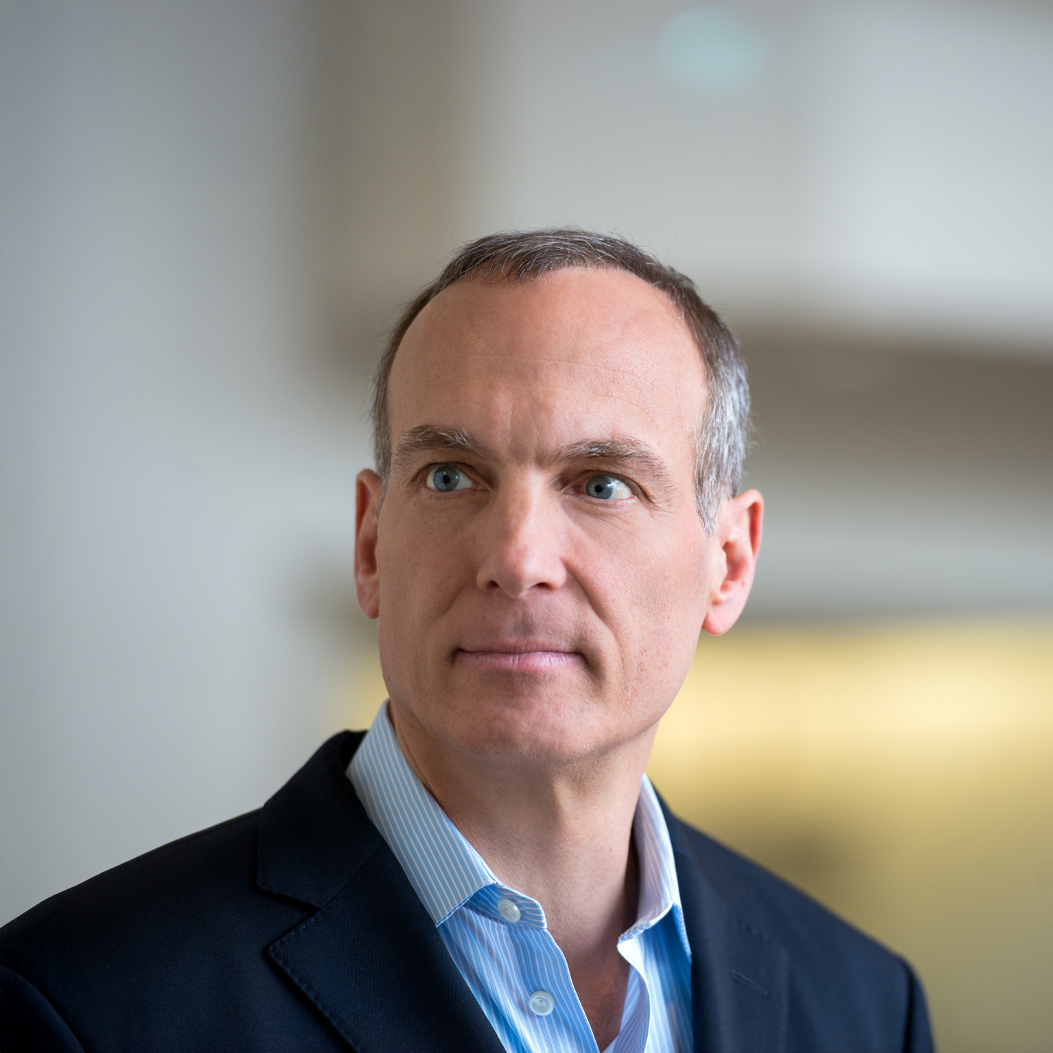 Glenn Fogel with short salt and pepper hair and blue eyes, wearing a dark blazer over a light collared shirt. He is looking slightly off to the side with a neutral, thoughtful expression against a softly blurred indoor background.