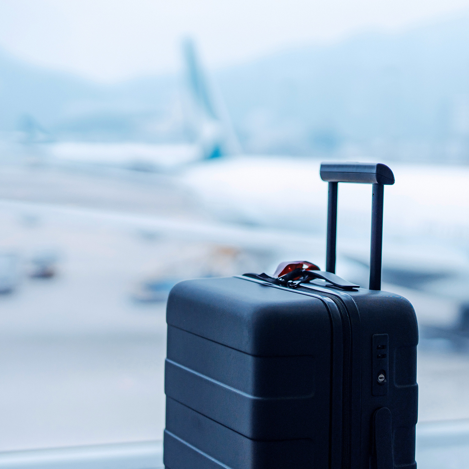 A black suitcase in front of the window at international airport terminal, with blurred airplane in the background.
