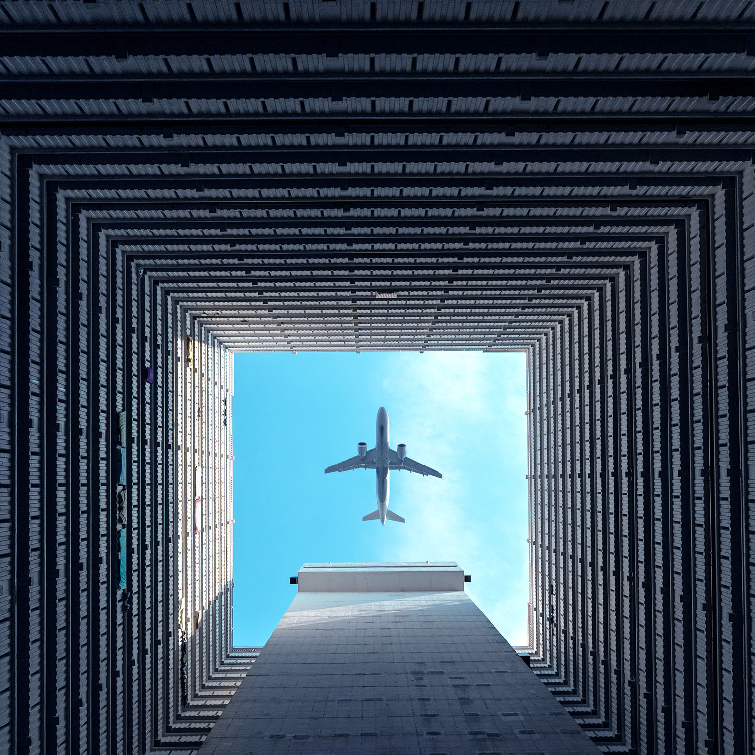 Airplane flying overhead, viewed through the open center of a high-rise building courtyard.