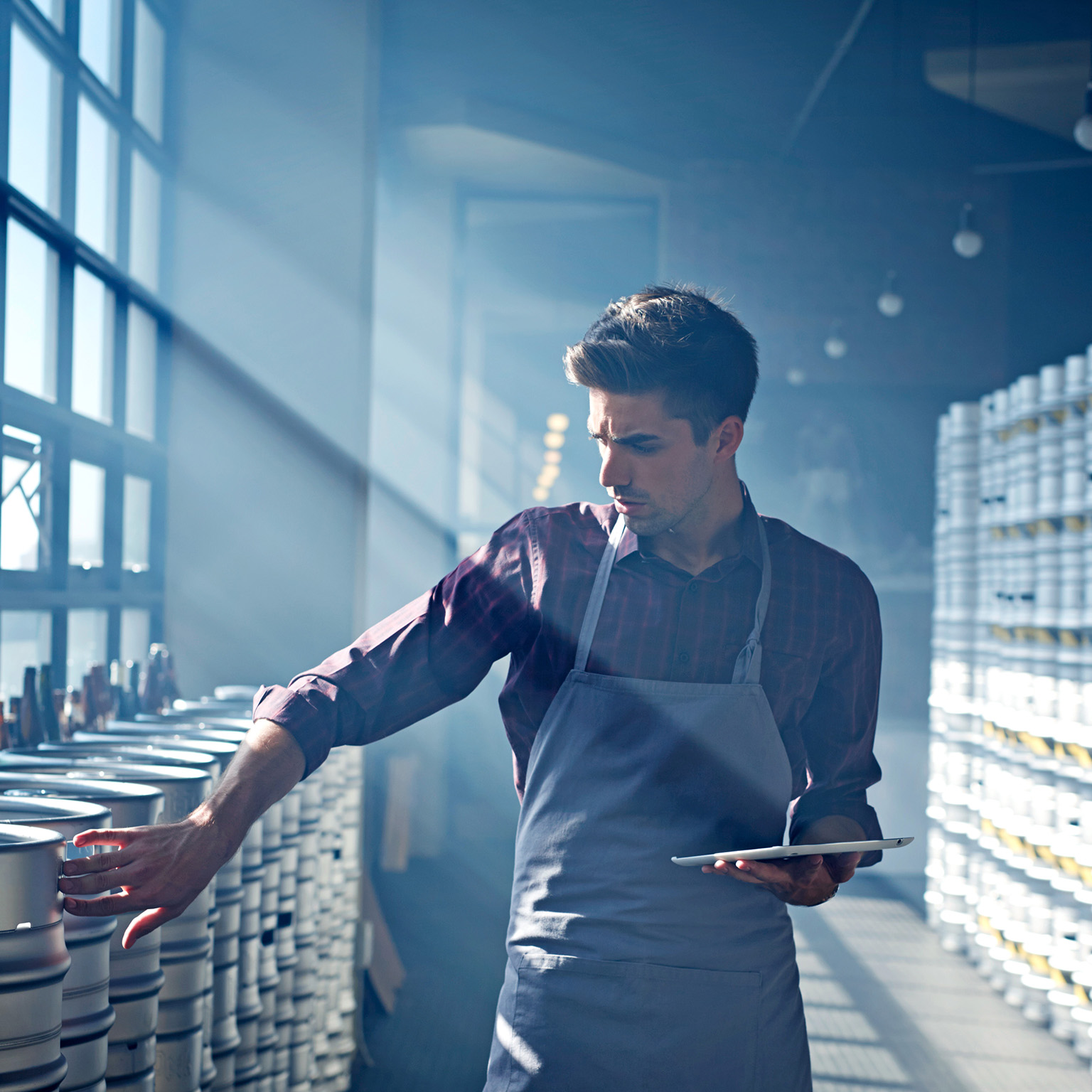 Quality control specialist in apron inspecting beer barrels in brewery, holding digital tablet in hands