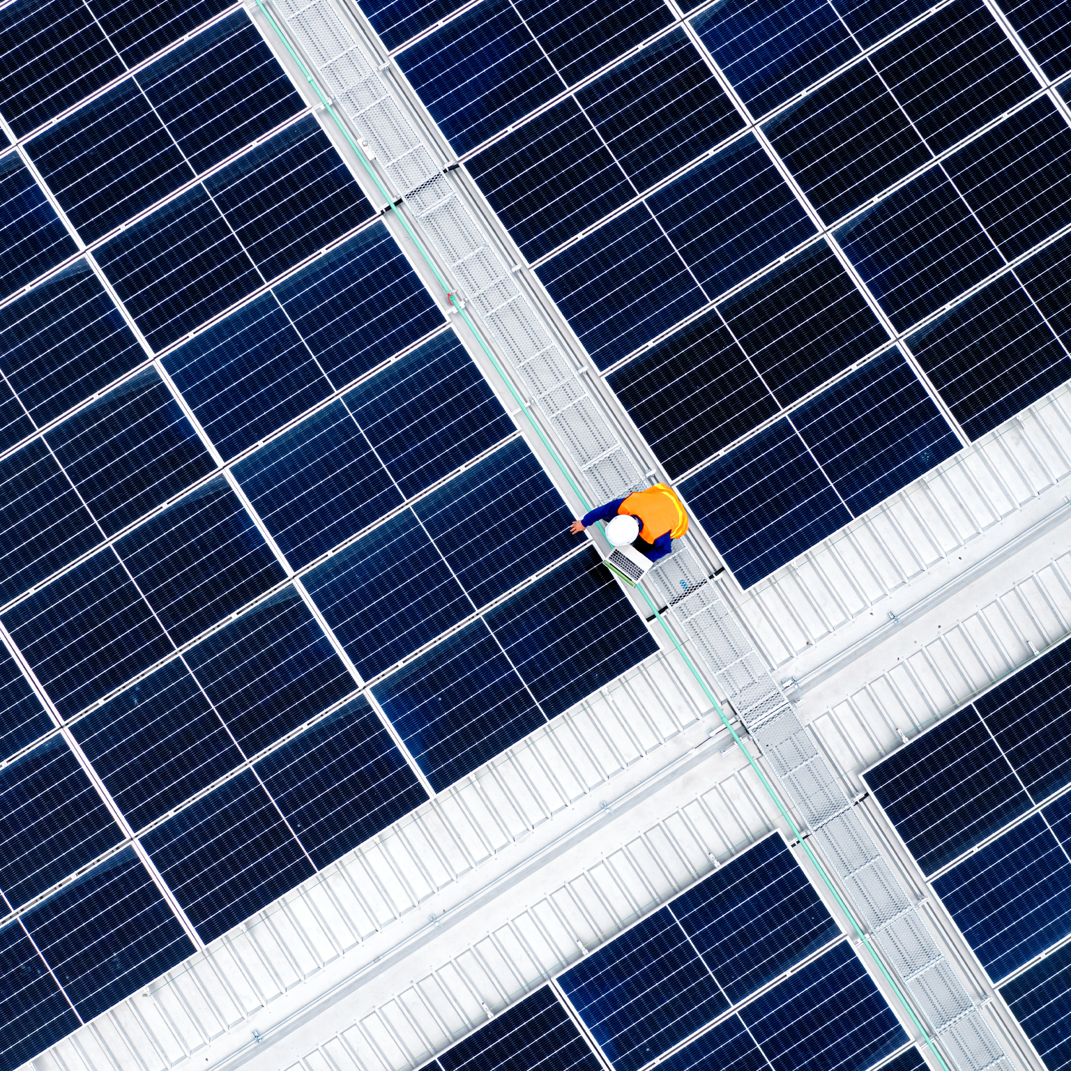 A vast array of dark blue solar panels arranged in a precise geometric grid, viewed from above. At the center, a worker in a bright orange safety vest walks along a maintenance pathway.