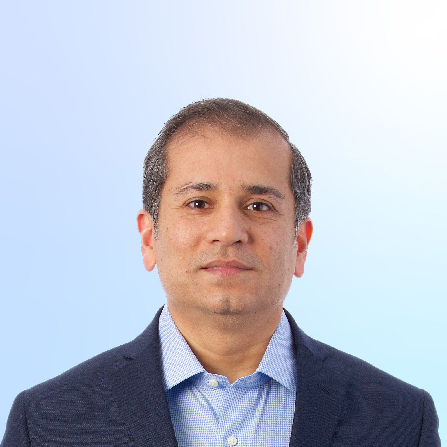 A head-and-shoulders portrait of a Shubham Singhal facing forward with a neutral expression. He is wearing a dark blazer over a light blue patterned dress shirt, set against a soft gradient background that transitions from light gray to pale blue.