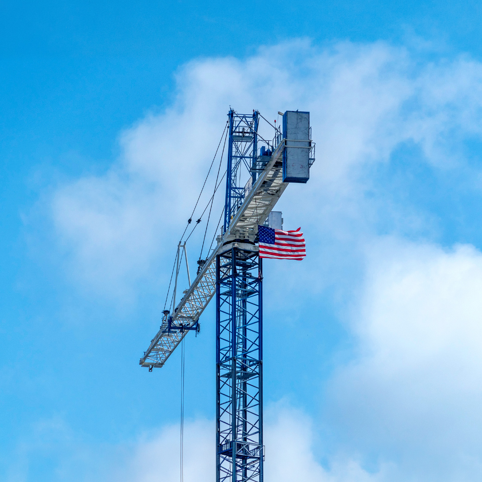 An American flag attached near the top of a tall construction crane waves in the wind. The crane rises against a cloudy blue sky, with its long arm extending outward.
