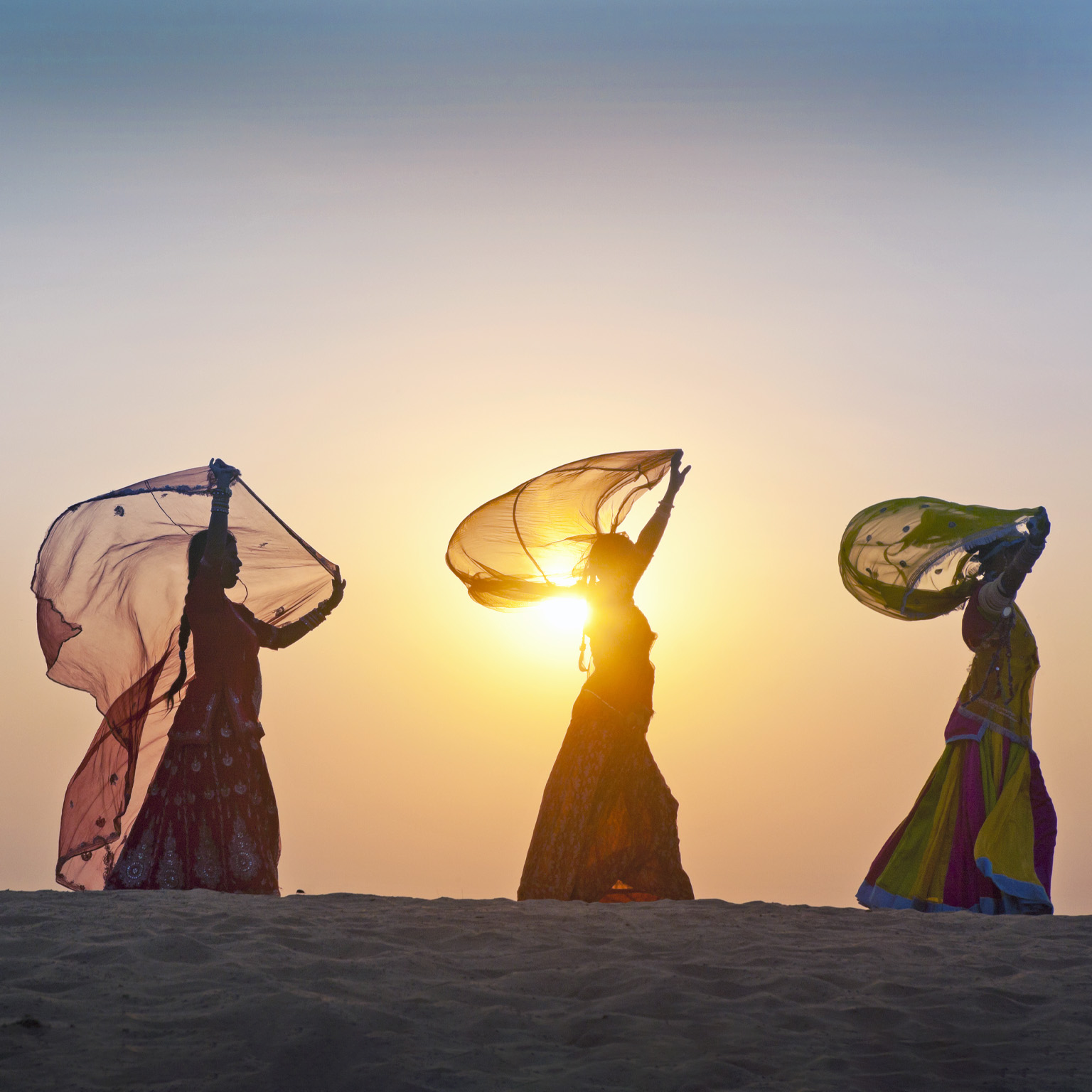 As the sun rises behind them, four Indian women dressed in traditional attire stroll along the crest of a desert dune, casting their figures in silhouette.