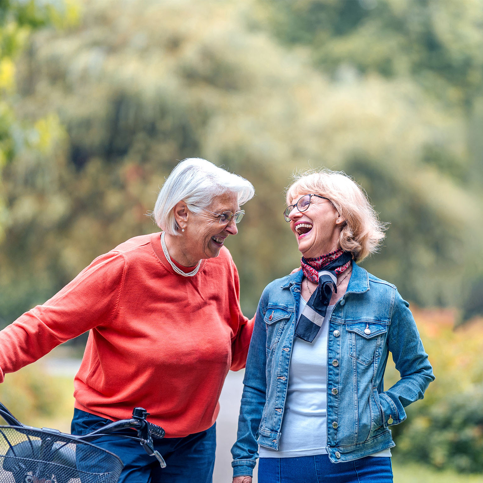 Two joyful senior women enjoy a leisurely walk in a lush park, one friend casually pushing a bicycle, both dressed in casual outdoor wear.
