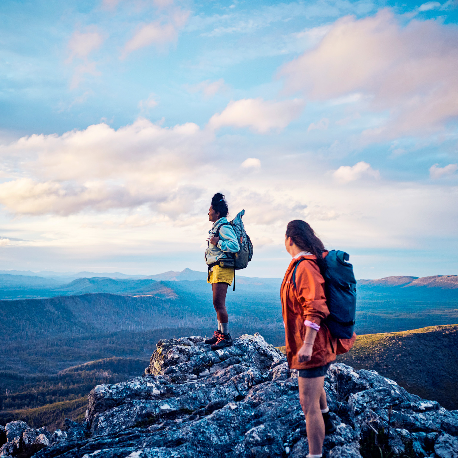 Two hikers stand on a rocky mountain peak, wearing backpacks and looking out over a vast, scenic landscape. The expansive view and soft light convey a sense of adventure, achievement, and connection with nature.