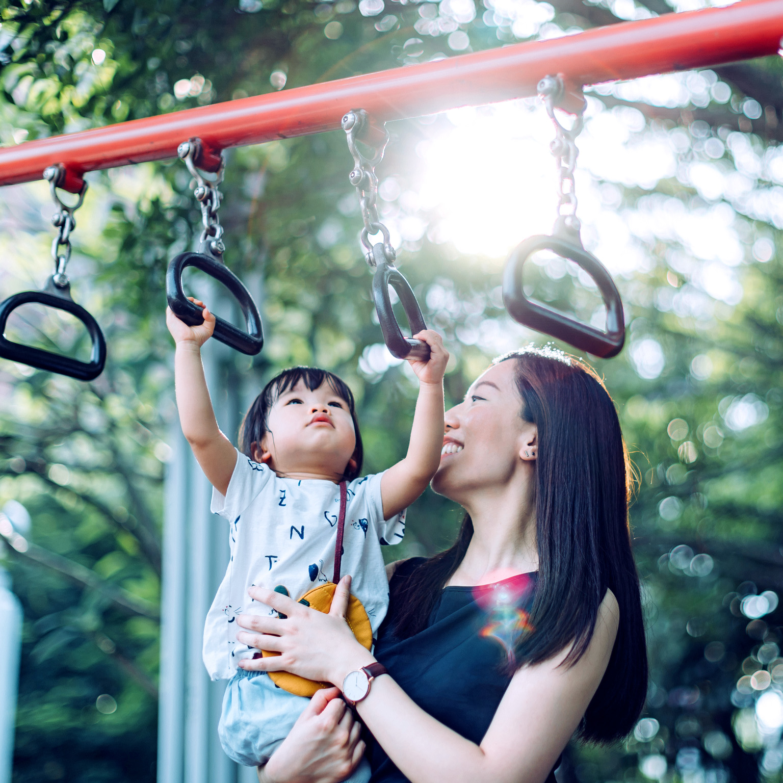 A mother smiles as she holds her young child up while the child grasps a monkey bar in an outdoor playground.