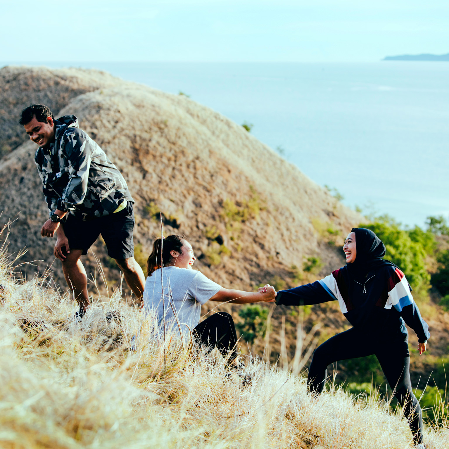 Three smiling friends, who are slightly heavyset, are climbing a grassy hill. One friend is assisting the other to stand up from a sitting position. The view from the top of the hill offers a peaceful ocean scene bathed in golden sunlight.