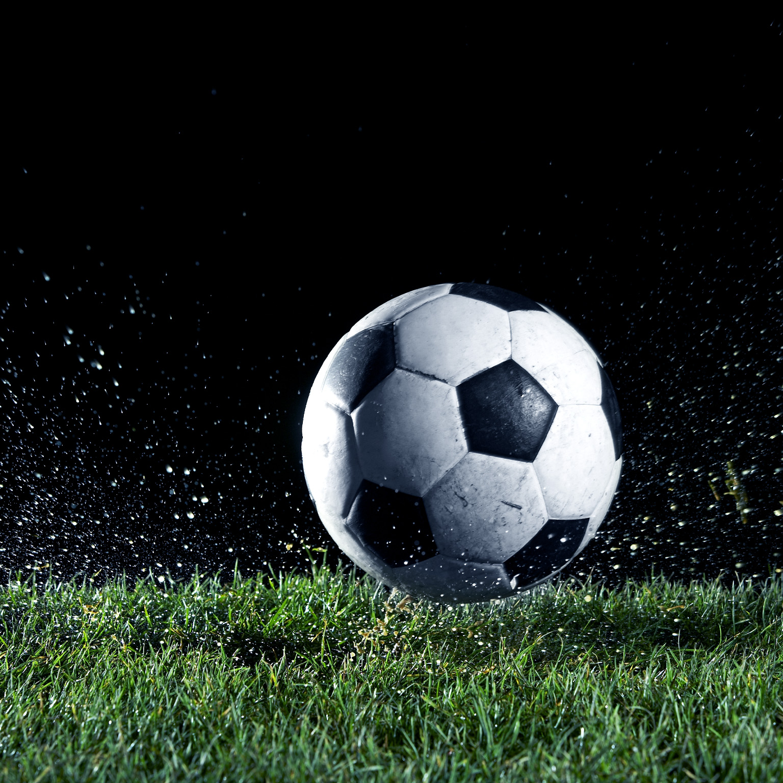 A black-and-white soccer ball sits on wet grass as water and turf spray outward, suggesting recent impact or motion. 