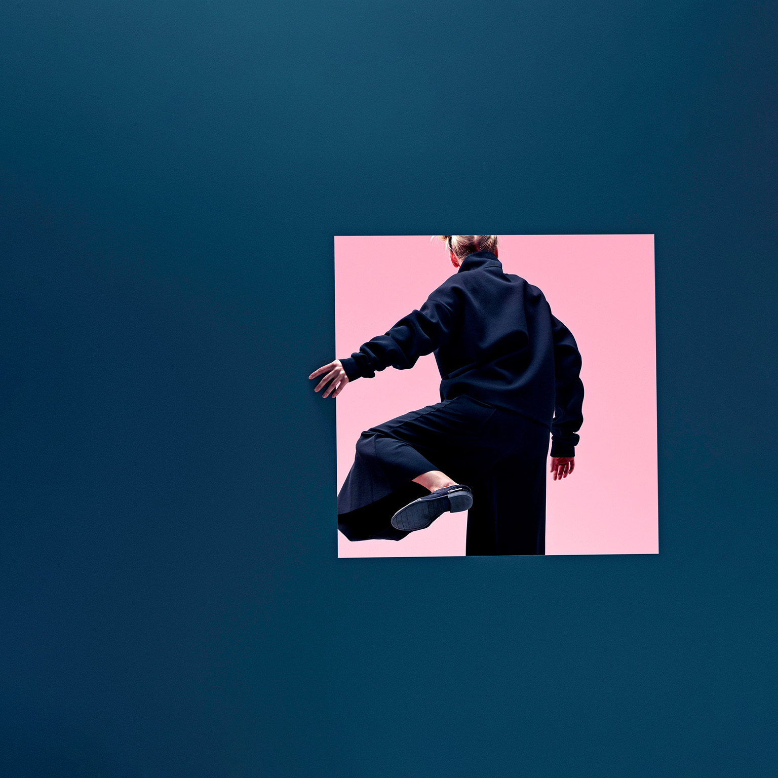 A woman in loose fashionable clothing stepping though a square opening half her height in a dark blue colored wall. The other side of the opening is bright and pink. 