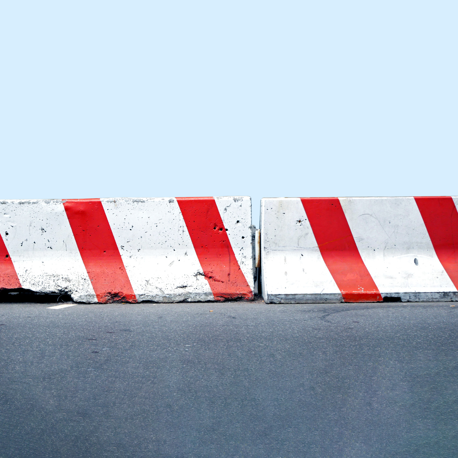 A pair of red and white concrete road barriers aligned on a street against a light blue background. 