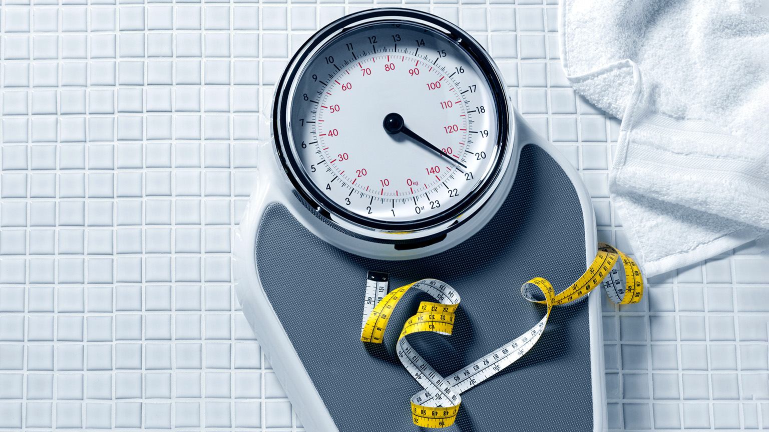 A bathroom scale with a large, round face sits on a tiled floor, next to a folded white towel. A yellow measuring tape is coiled on the scale's platform.