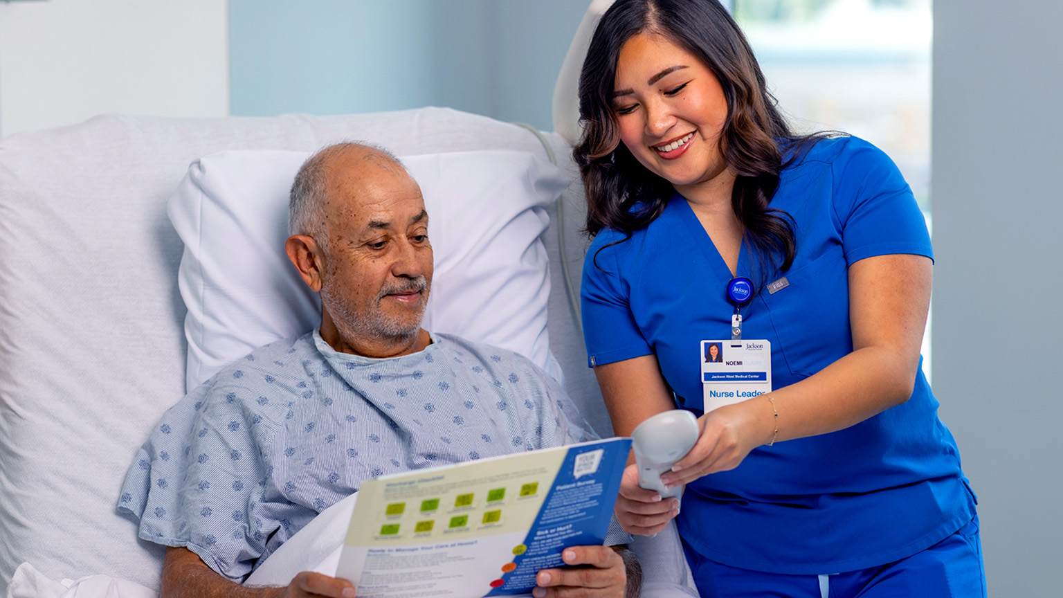 Nurse in blue scrubs consulting with patient in hospital bed, reviewing educational materials