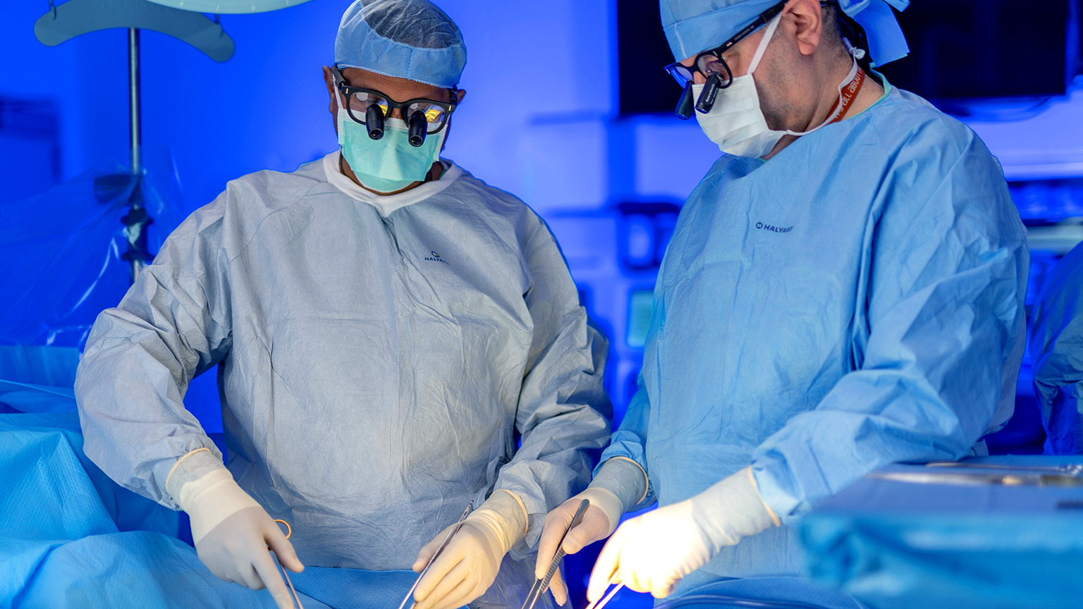 Two surgeons in surgical gowns and masks performing a procedure in an operating room under blue surgical lighting