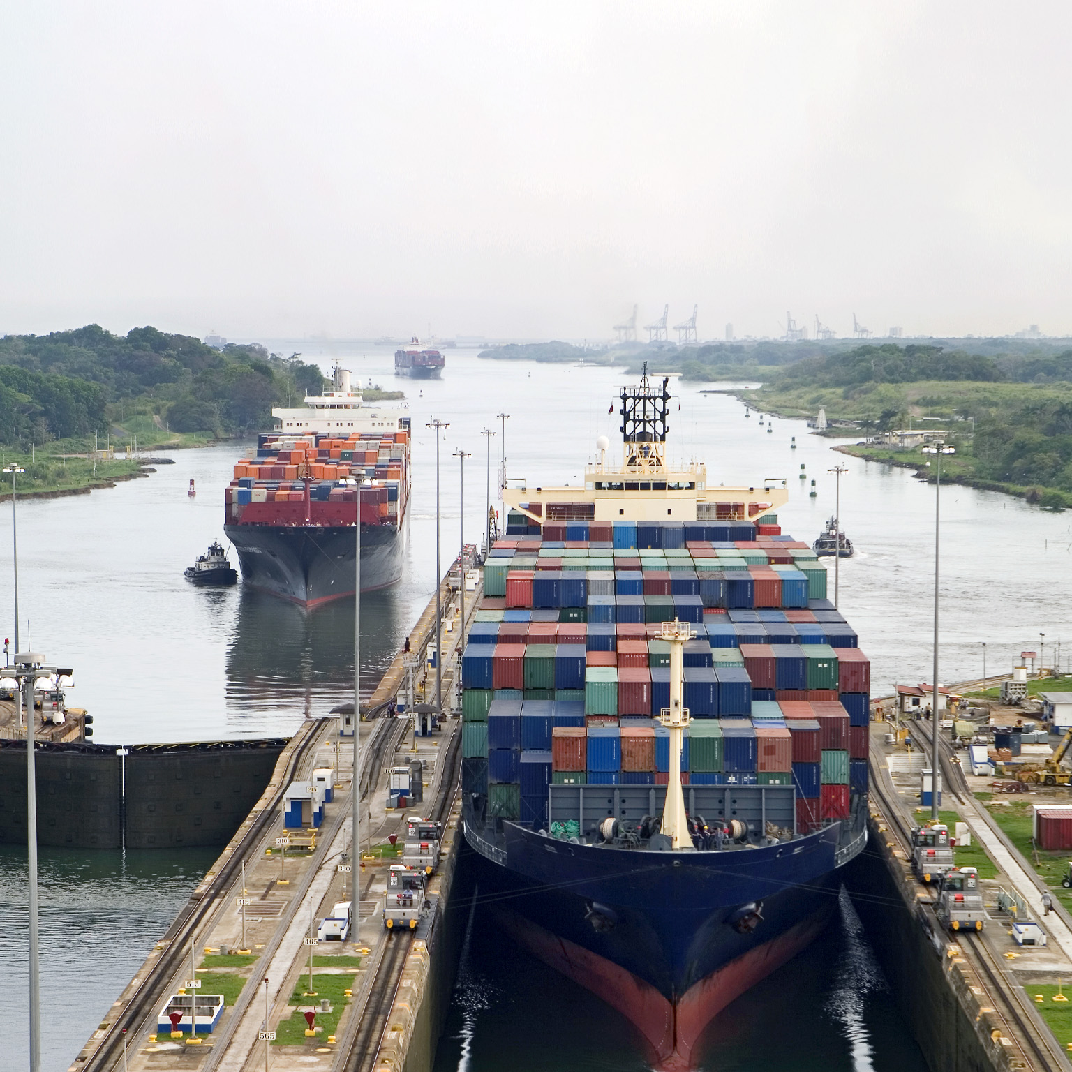 Cargo ships assisted by tugboats, entering the Panama Canal at Gatun Locks on the Atlantic side. The ships are fully loaded with cargo heading west towards the Pacific.