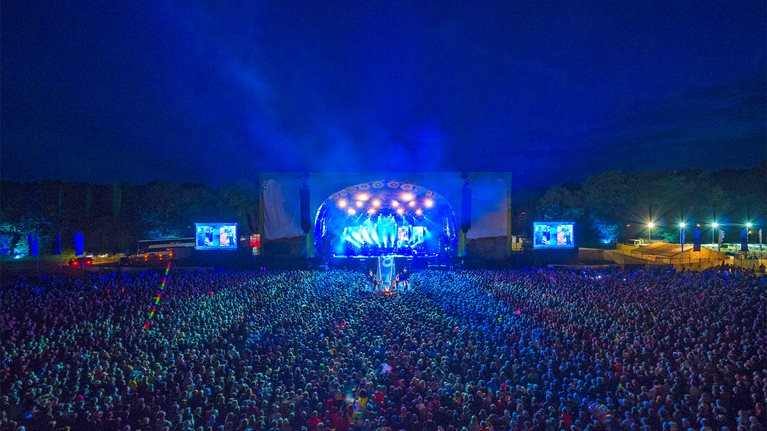 A large crowd watching a band perform at night during a summer music festival in the UK.