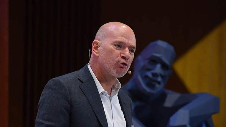 Andy McAfee speaking on stage with a smiling blue statue in a relaxed pose behind him.