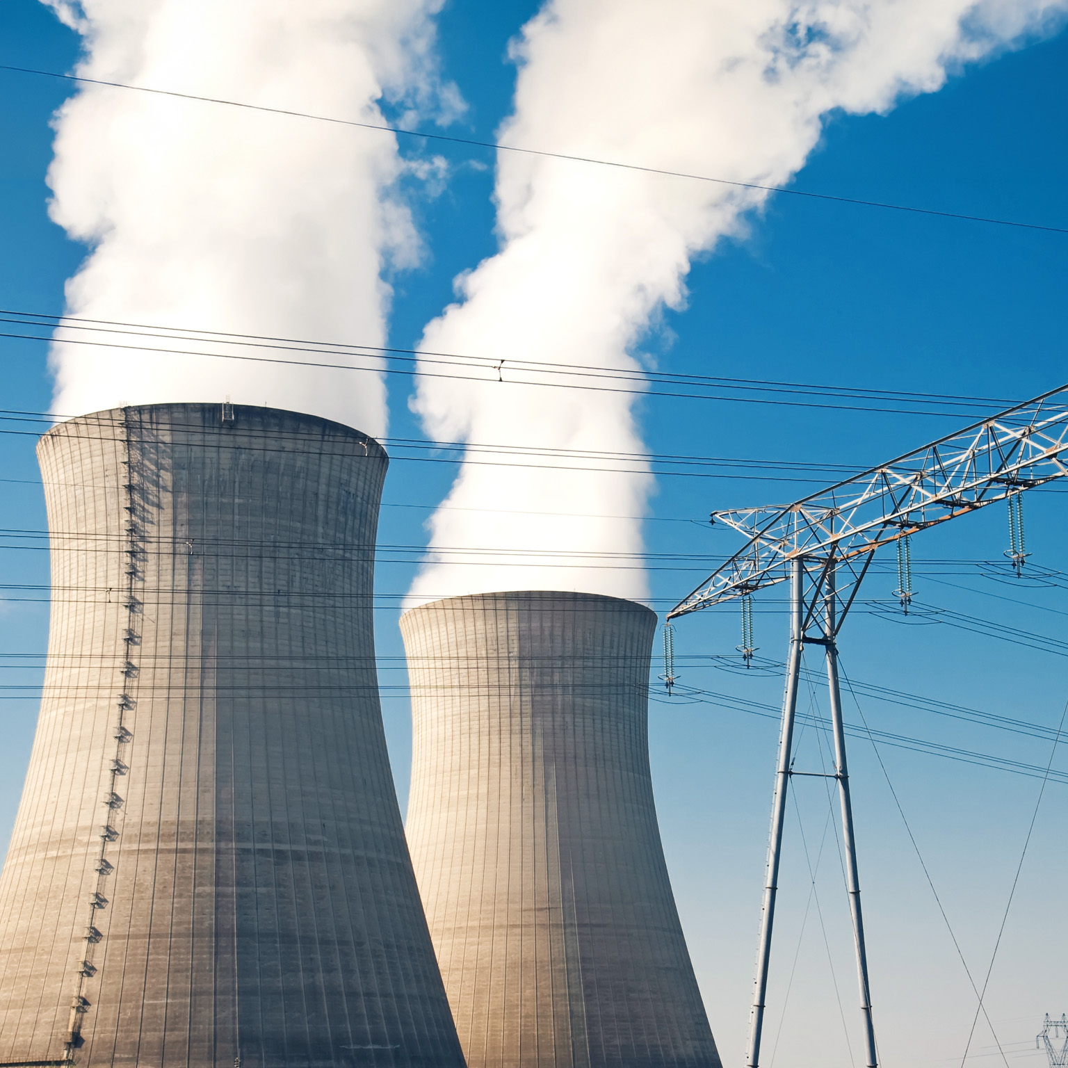 Two large cooling towers at a power plant releasing thick white steam into a clear blue sky, with high-voltage transmission lines and electrical towers in the foreground.