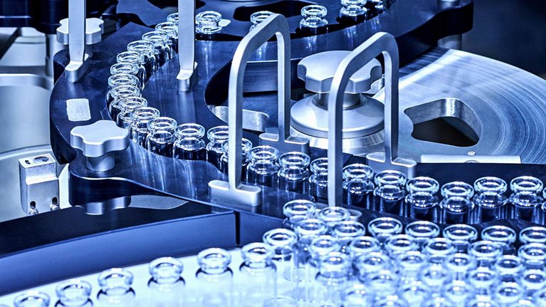 A pharmaceutical production line with rows of small glass vials moving through a complex stainless-steel machine.