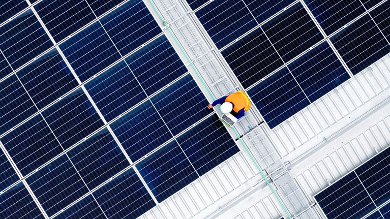 A vast array of dark blue solar panels arranged in a precise geometric grid, viewed from above. At the center, a worker in a bright orange safety vest walks along a maintenance pathway.