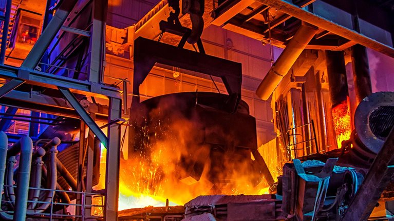 The interior of a large industrial steel plant, where molten metal glows bright orange amid heavy machinery and towering metal structures.