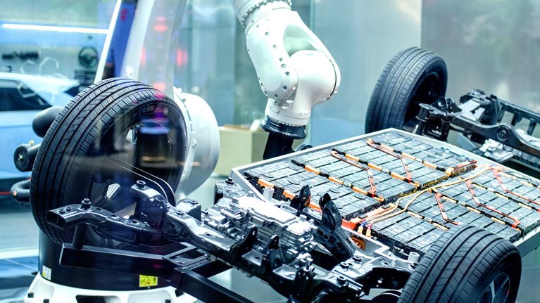 The exposed chassis and battery platform of an electric vehicle surrounded by robotic arms in a high-tech manufacturing or display environment.