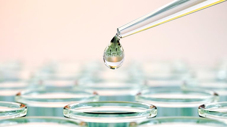 A close-up of a laboratory pipette releasing a droplet of liquid above a row of clear test tubes filled with fluid.