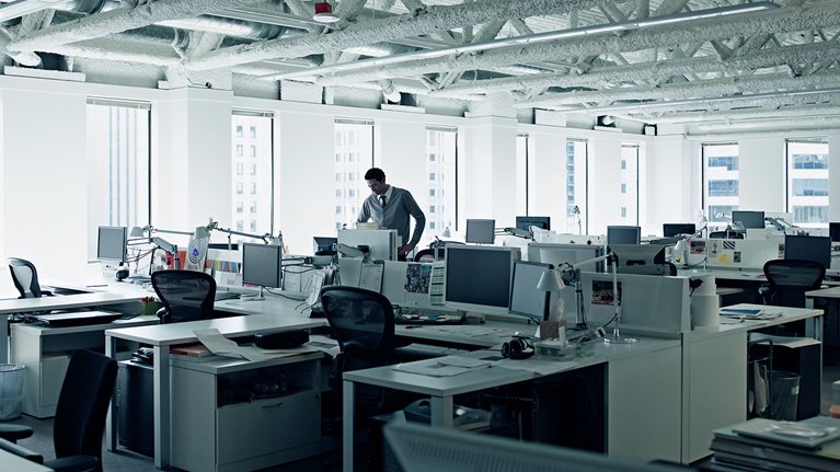 A businessman standing at desk in an empty office looking down at his computer monitor with his arms on his hips.