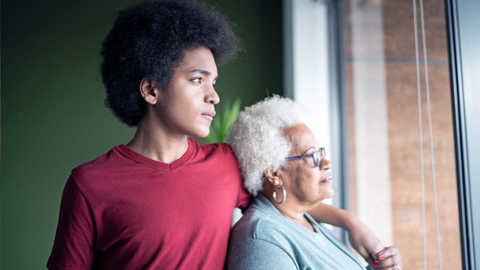 A young African American man stands beside his grandmother, his arm resting gently on her shoulder as they both gaze thoughtfully out of a window. 