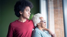 A young African American man stands beside his grandmother, his arm resting gently on her shoulder as they both gaze thoughtfully out of a window.